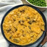 A dark blue serving dish filled with mushroom chicken in mustard sauce. There is a dish of peas in the background.