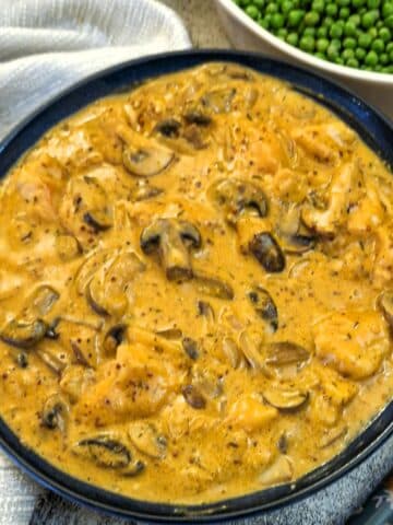 A dark blue serving dish filled with mushroom chicken in mustard sauce. There is a dish of peas in the background.