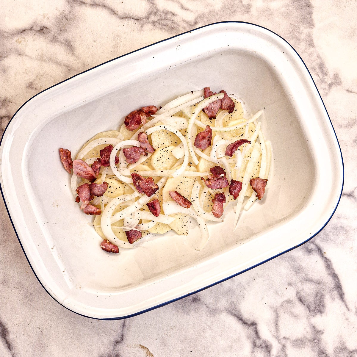 A layer of potatoes, onions and bacon in a white enamel baking dish.