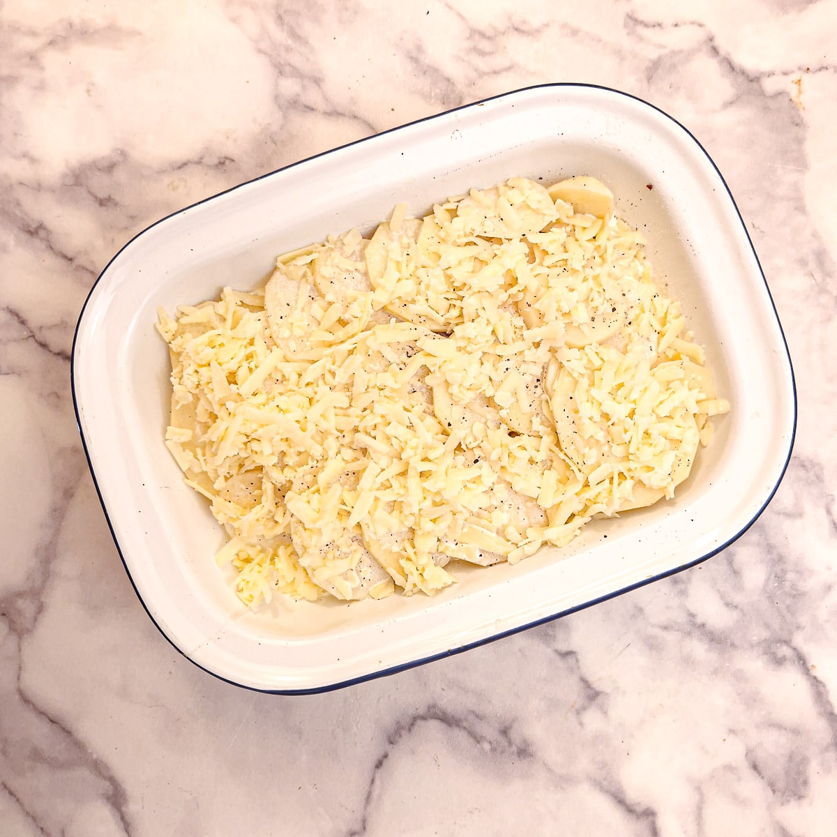 A fully assembled potato bake covered in grated cheese in a white enamel baking dish.