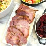 Slices of gammon on a white serving platter surrounded by dishes of salads.