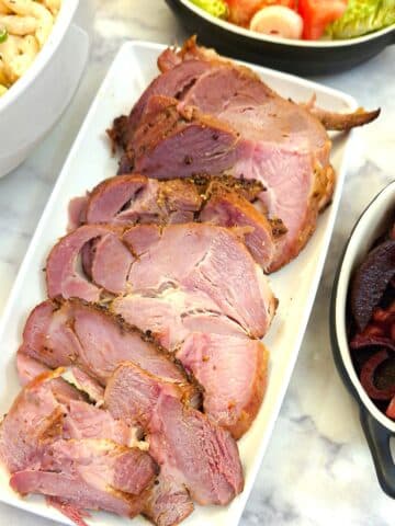 Slices of gammon on a white serving platter surrounded by dishes of salads.