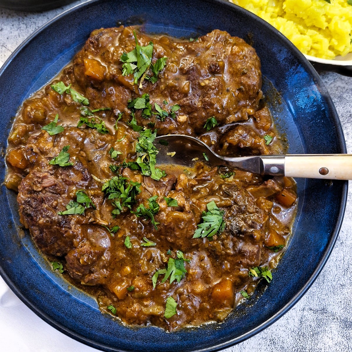 A blue serving dish with beef osso buco garnished with parsley.