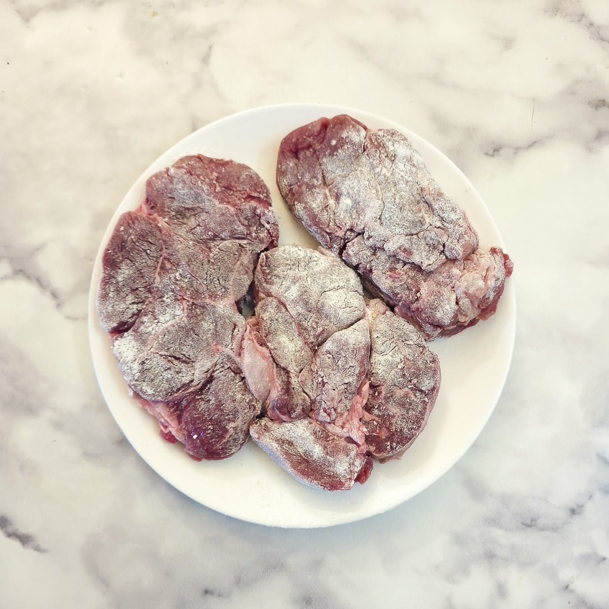 Three pieces of beef shin which have been seasoned and coated with flour on a white plate.