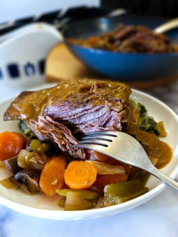 A piece of slow cooker beef short rib on a dinner plate with vegetables. The beef is being pulled apart with a fork. There is a helping of thick gravy over the beef.