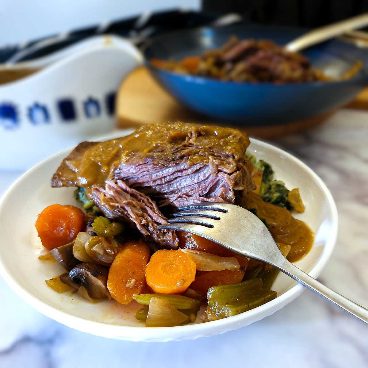 A piece of slow cooker beef short rib on a dinner plate with vegetables. The beef is being pulled apart with a fork. There is a helping of thick gravy over the beef.