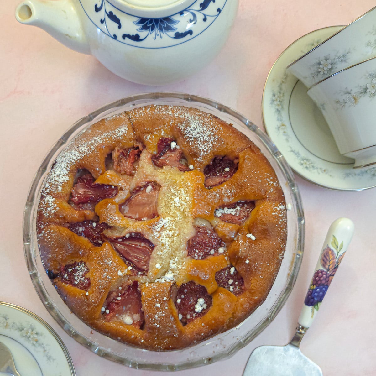 Overhead shot of a strawberry mascarpone cake on a glass platter surrounded by a teapot and teacups.