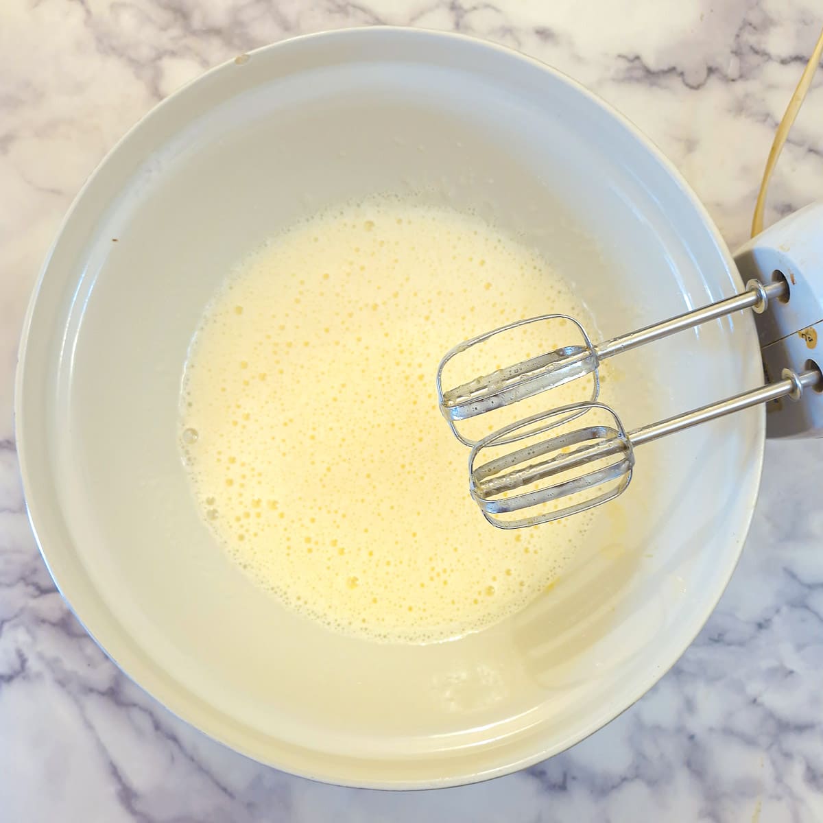 Eggs and sugar whisked together in a mixing bowl.  There is an electric beater on resting on the side of the mixing bowl.