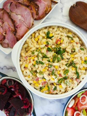 Overhead shot of a creamy crab and roasted pepper pasta salad. The salad is in a white bowl and is surrounded by dishes of other salads and a platter of cold meat.