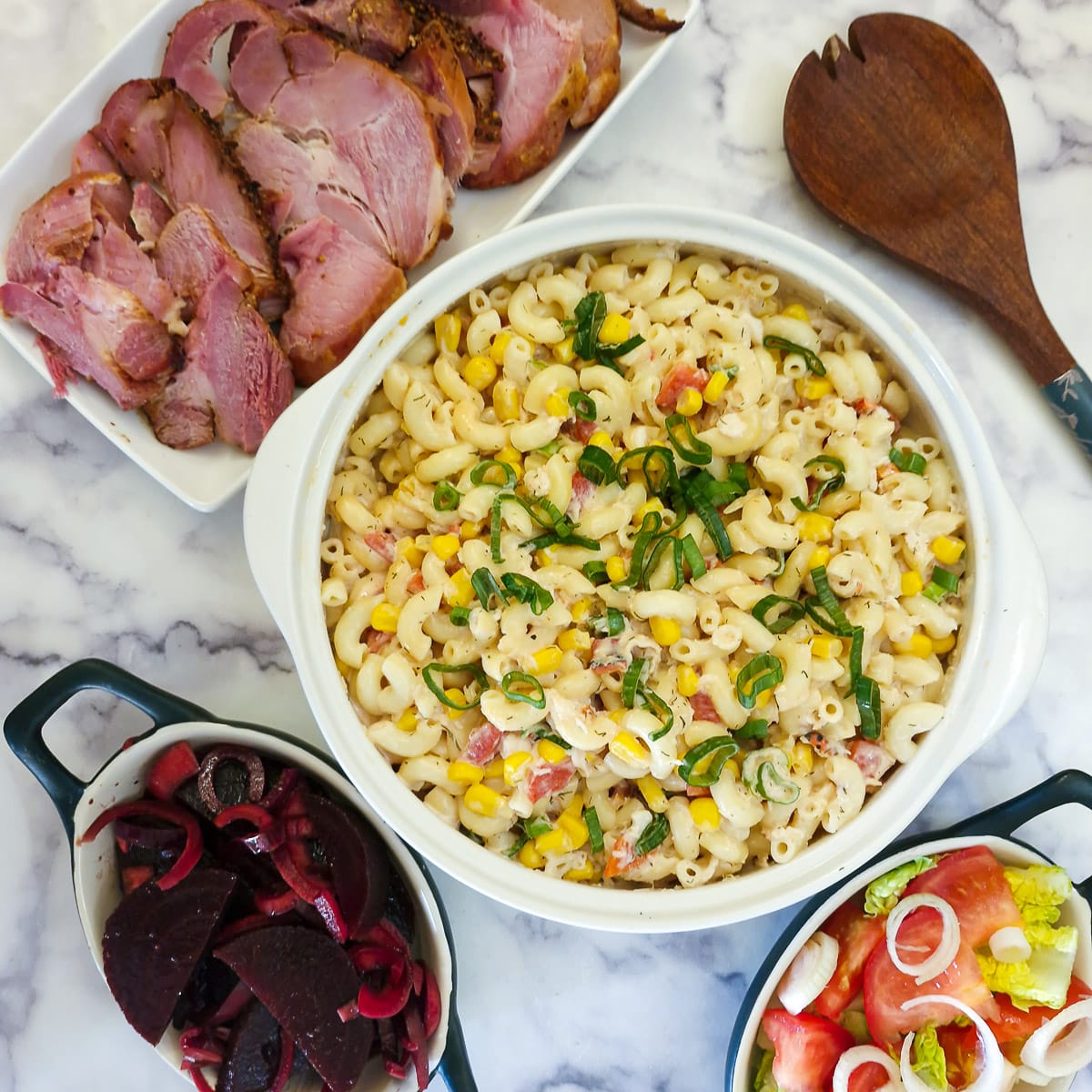 Overhead shot of a creamy crab and roasted pepper pasta salad. The salad is in a white bowl and is surrounded by dishes of other salads and a platter of cold meat.