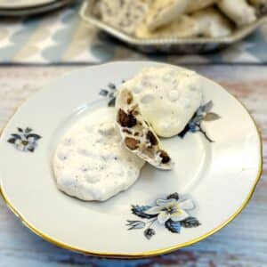 Two crispy meringue nut cookies on a cake plate with one broken in half to show the choclate chips and nuts on the inside.