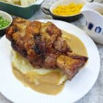 A crispy pork hock on a pile of mashed potatoes, surrounded with gravy on a white plate.