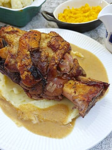 A crispy pork hock on a pile of mashed potatoes, surrounded with gravy on a white plate.