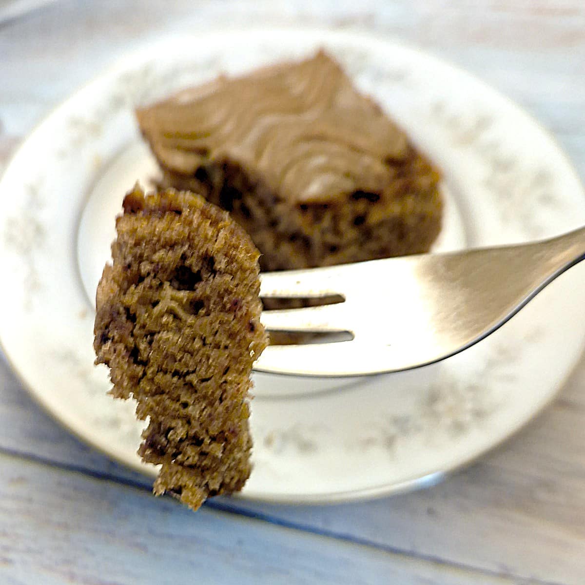 A piece of toffee cake on a fork with a blurred s;lice of cake in the background.