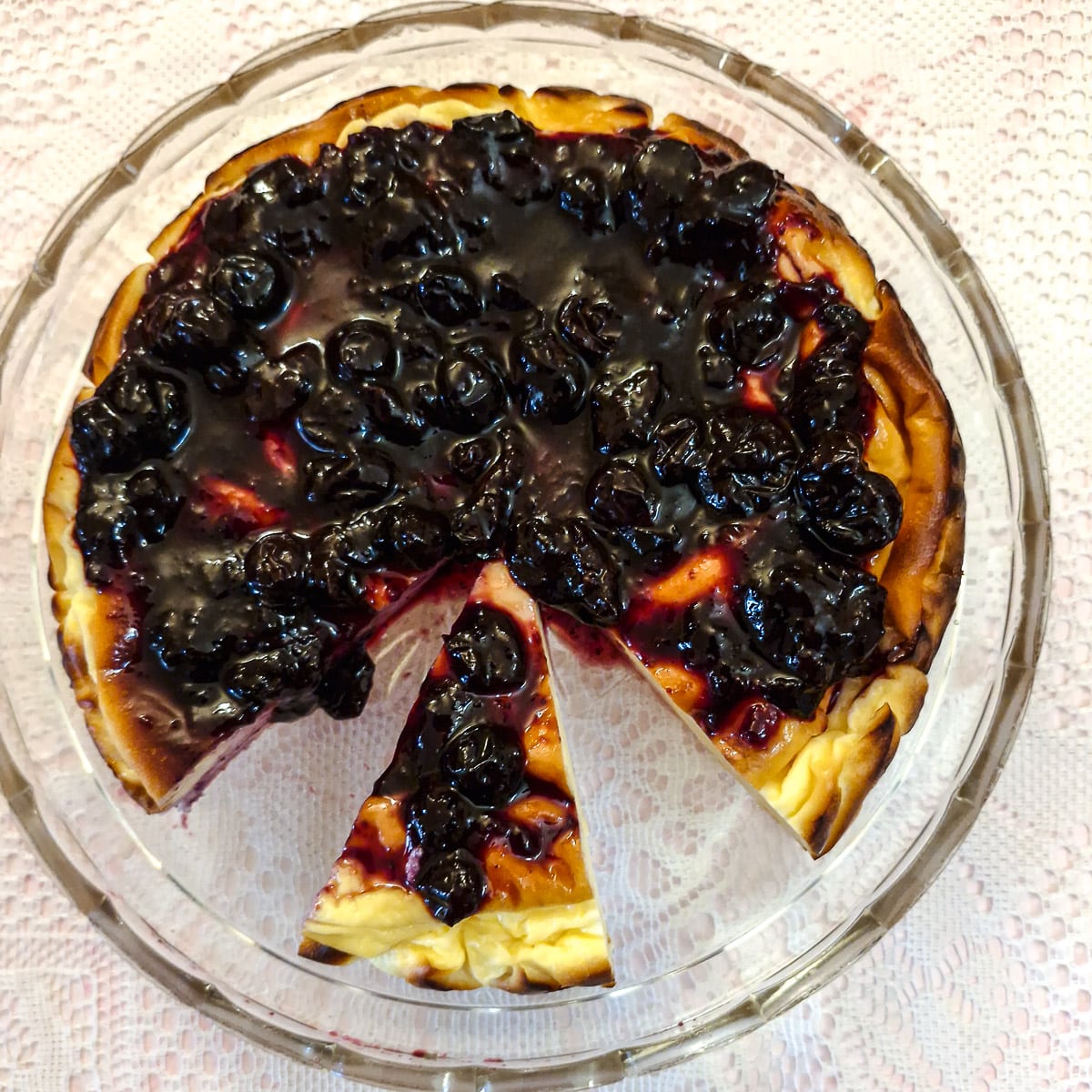Overhead shot of a yoghurt chesecake on a glass plate, topped with blueberry compote, with a slice offset.