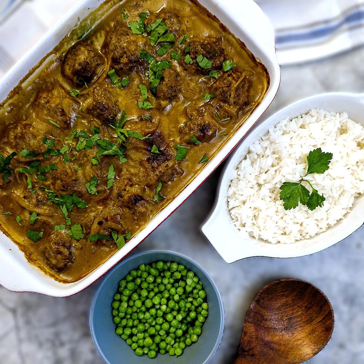 A dish of meatballs in curried onion sauce on a table next to a bowl of peas and a dish of rice.