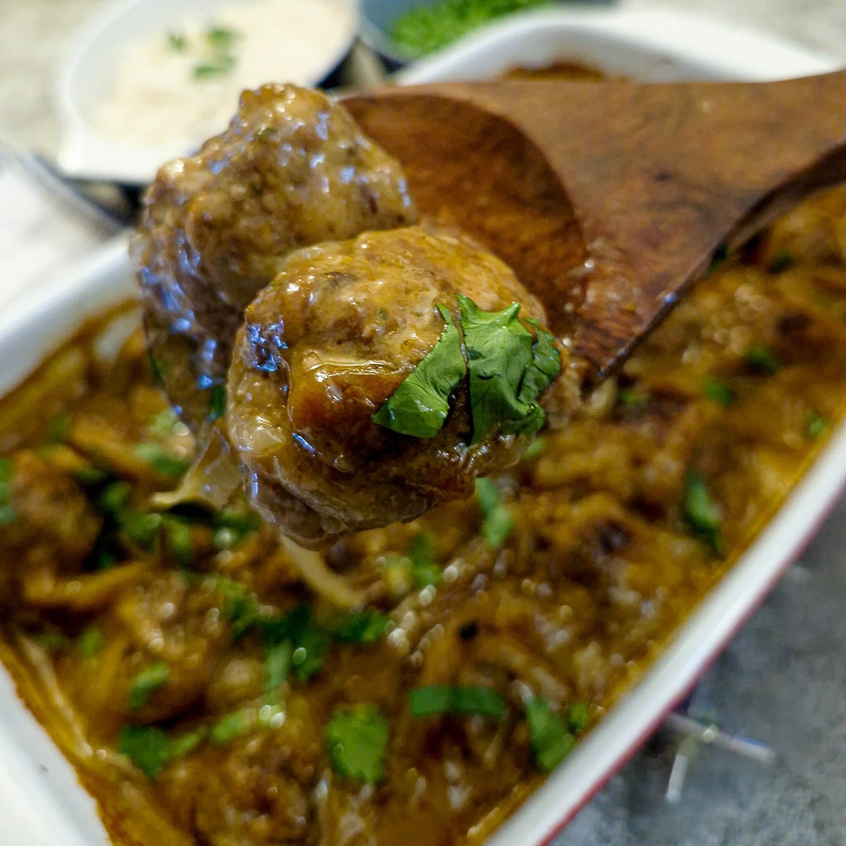 Two South African-style curried meatballs on a wooden spoon held over the cassereole dish.