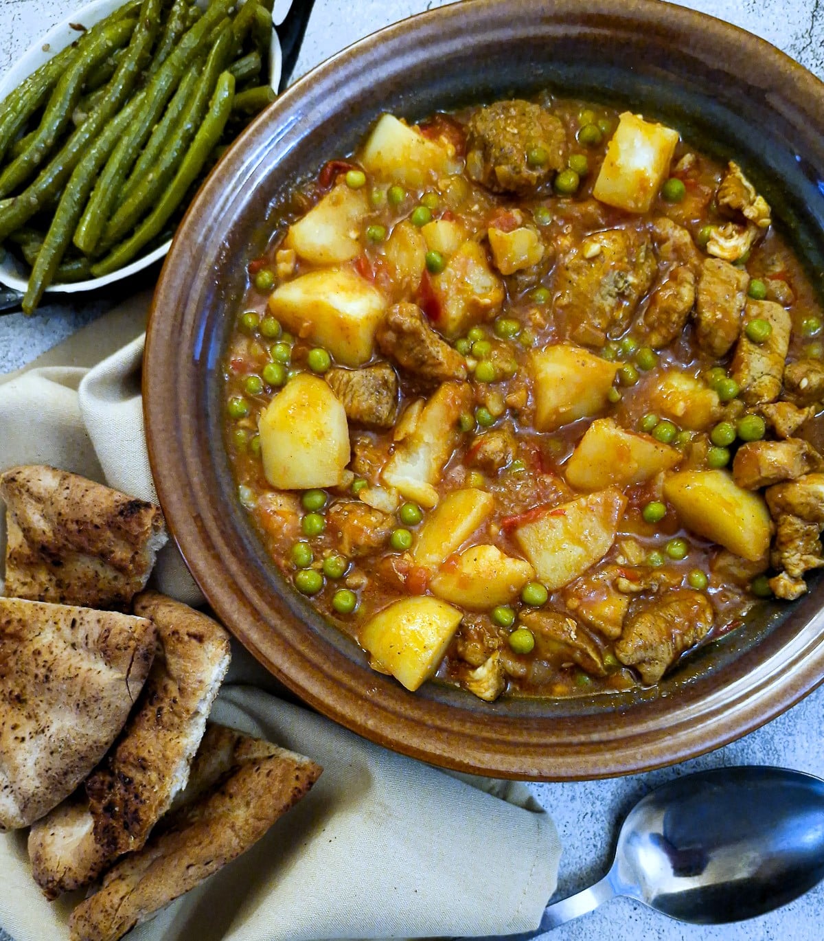 Chicken aloo matar in a serving dish with dishes of naan bread and green beans on the side.