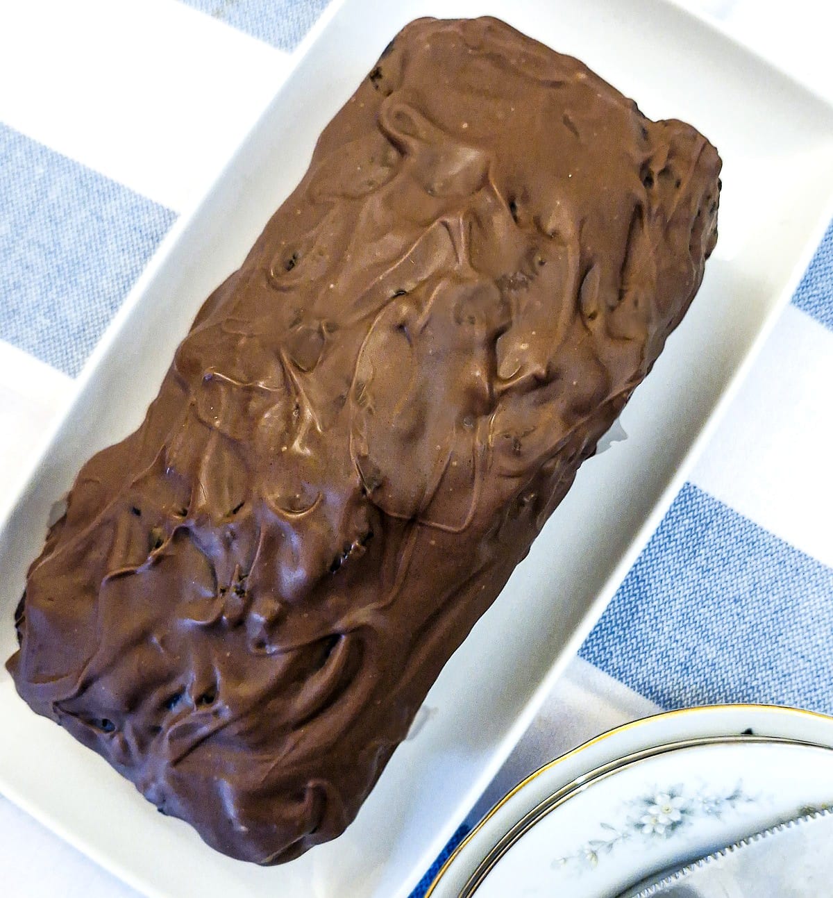 A chocolate coated cake on a white serving platter, on a blue and white striped tablecloth.