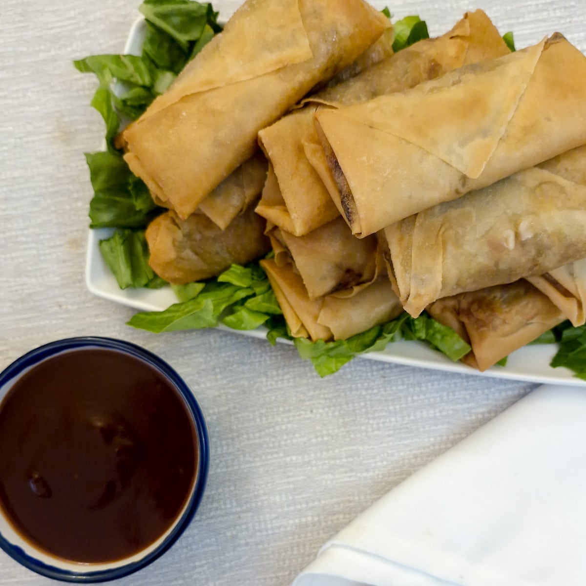A pile of crispy prawn spring rolls on a plate next to  bowl of dipping sauce.