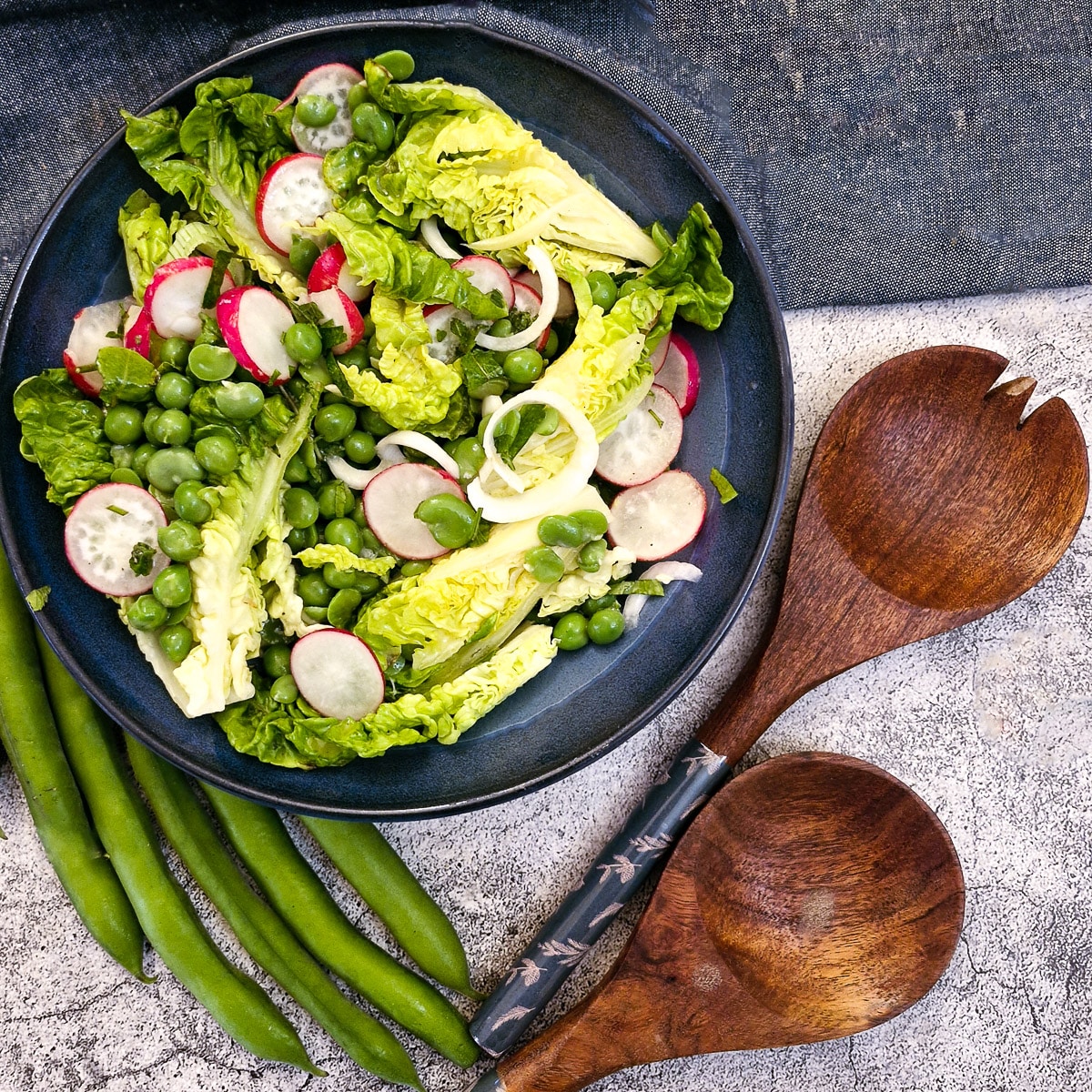 An assembled pea and broad bean salad in a blue serving dish with wooden salad servers in the foreground.