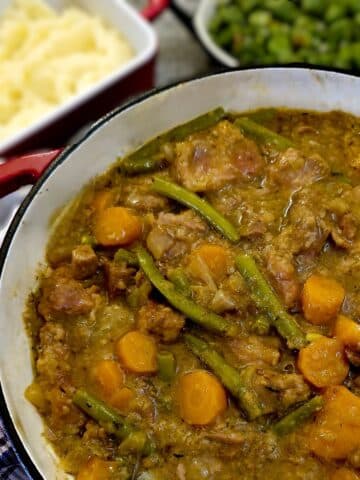 A dish of pork and apple casserole on a table with mashed potatoes an broad beans.