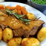 Sliced pot roast of brisket on a serving platter, garnished with rosemary, with potatoes and carrots on the side.