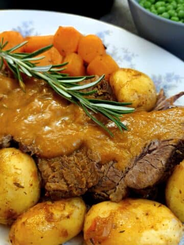 Sliced pot roast of brisket on a serving platter, garnished with rosemary, with potatoes and carrots on the side.