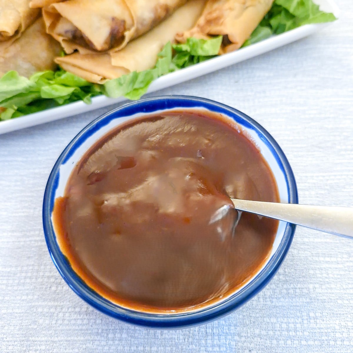 A small bowl of sweet and sour dipping sauce with a spoon.  There is a plate of spring rolls in the background.