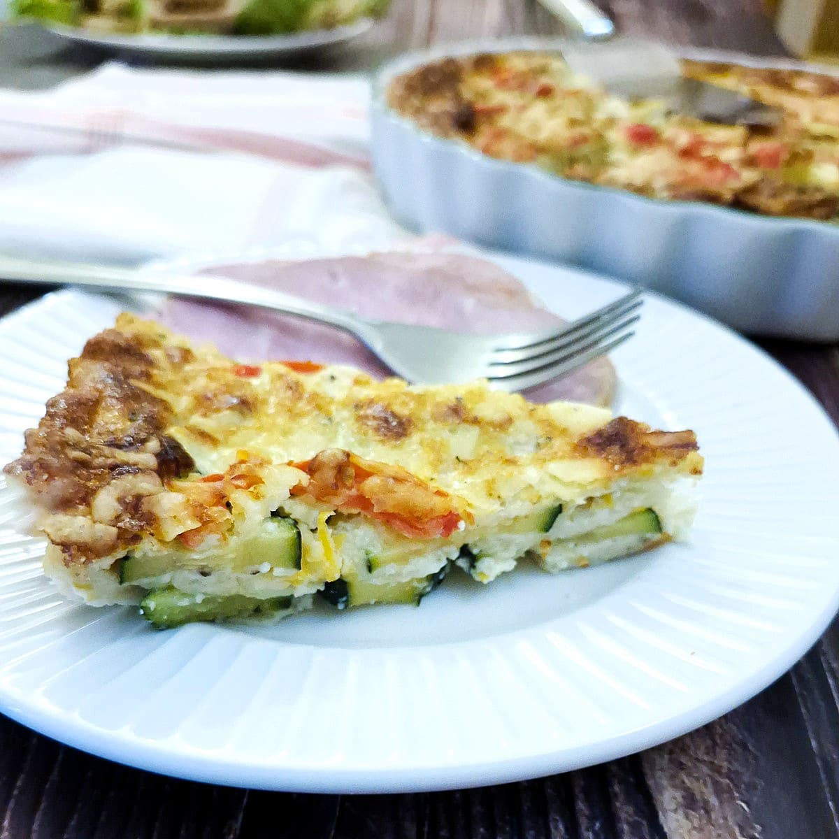 Closeup of a slice of courgette and carrot quiche on a white plate with a slice of gammon and a fork.