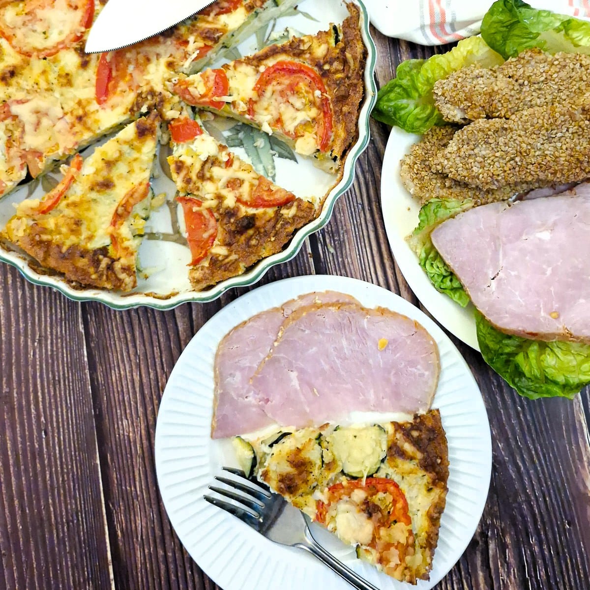 A slice of crustless zucchini quiche on a smal white plate with a slice of gammon.  The zucchini quich is in a baking dish in the background.