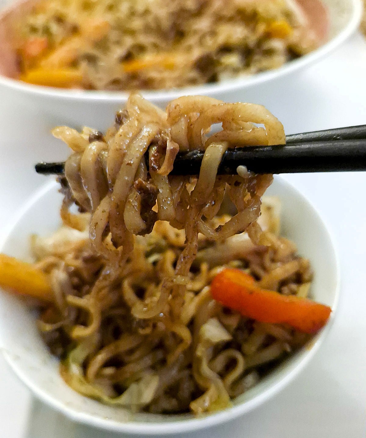 Noodles with beef and vegetables held in a pair of chopsticks over a bowl of chow mein.