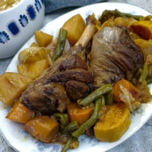 Two braised lamb shanks and vegetables on a serving platter with a dish of gravy in the background.