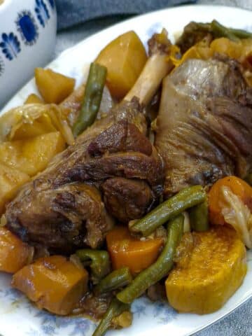Two braised lamb shanks and vegetables on a serving platter with a dish of gravy in the background.