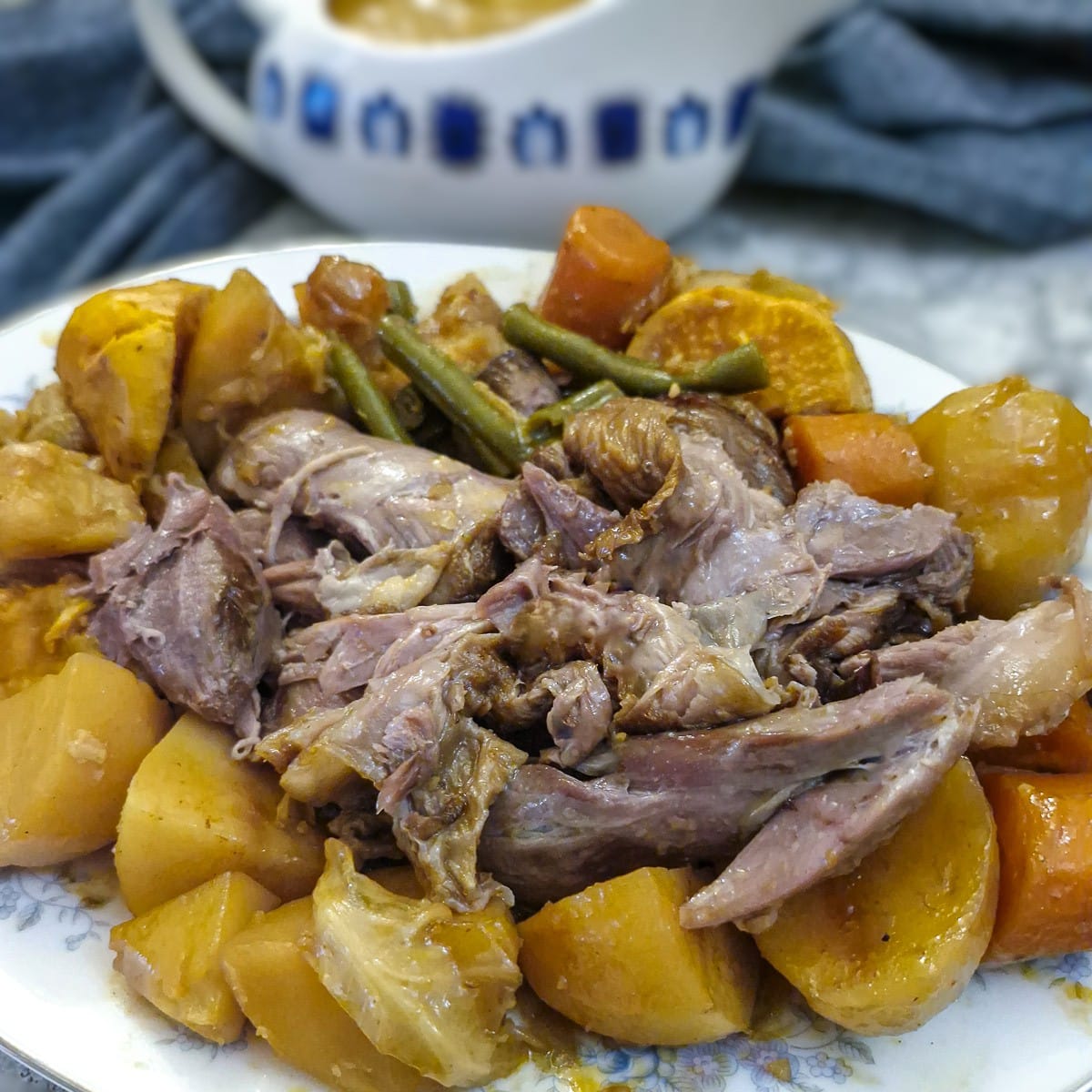 Shredded lamb shanks surrounded by vegetables on a serving platter. There is a jug of gravy in the background.