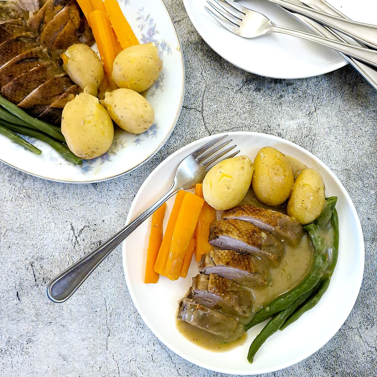 Overhead shot of sliced duck breasts and vegetables on a plate with the serving platter of duck and vegetables in the background.