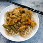 A white plate with garlic cashew chicken on a bed of noodles, with a pair of black chopsticks on the side.