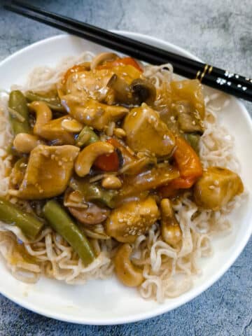 A white plate with garlic cashew chicken on a bed of noodles, with a pair of black chopsticks on the side.