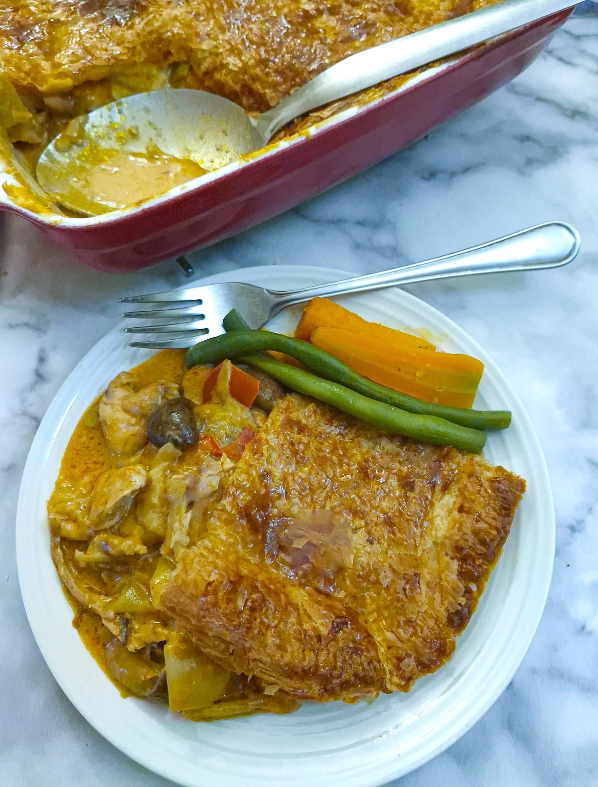 A helping of leftover chicken paprika pie on a plate with beans and carrots. A red serving dish is in the background.