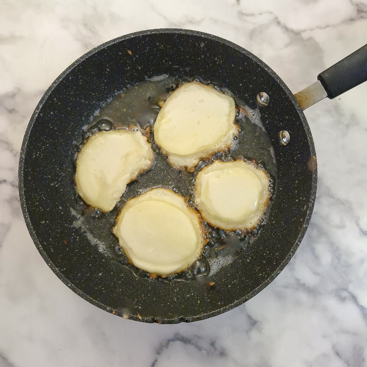 4 battered potato scallops being browned in hot oil in a frying pan.