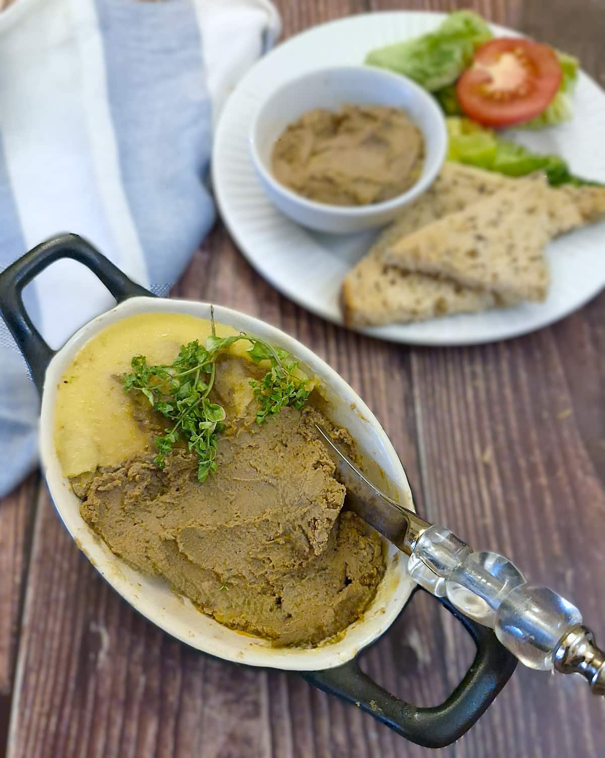 A small dish of pate on a table with a butter knife. Some of the melted butter seal has been removed and there is a plate of bread in the background.