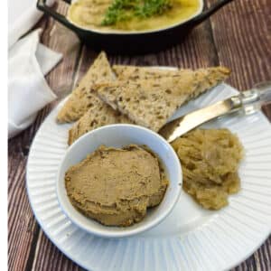 A small white dish of duck liver pate on a white plate with a spoonful of onion marmalade on the side and a few pieces of crusty brown toast.