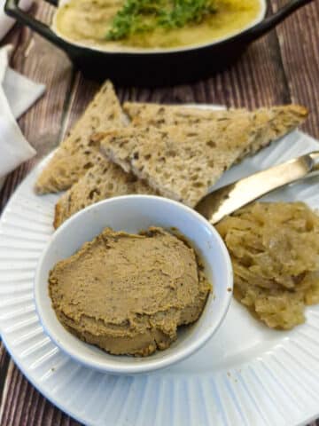 A small white dish of duck liver pate on a white plate with a spoonful of onion marmalade on the side and a few pieces of crusty brown toast.