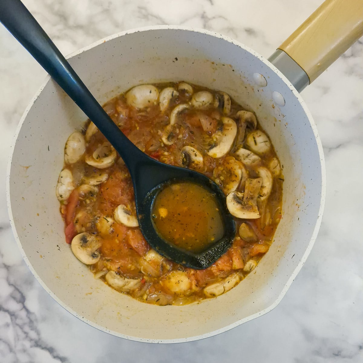 A saucepan of partly cooked onions, tomatoes and mushrooms, with a large spoon showing the amount of liquid in the pan.