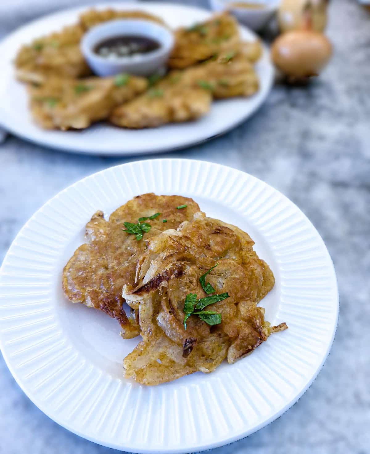 Close up of 2 onion fritters on small white plate with a larger serving plate in the background.