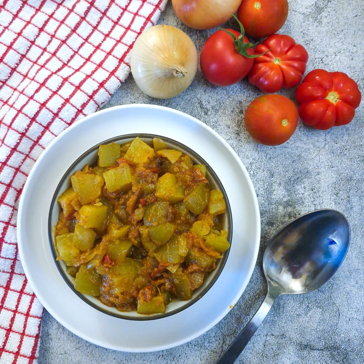 A dish of easy marrow curry in a white bowl along side a large serving spoon. There is a red and white checked cloth on the table.