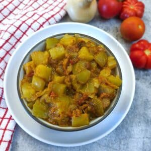 A white serving dish of curried marrow with tomatoes and onions. There are tomatos and onions on the table next to the dish.