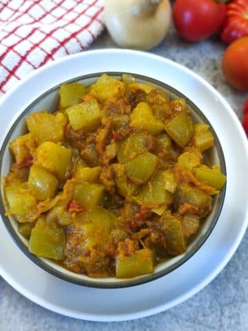 A white serving dish of curried marrow with tomatoes and onions. There are tomatos and onions on the table next to the dish.