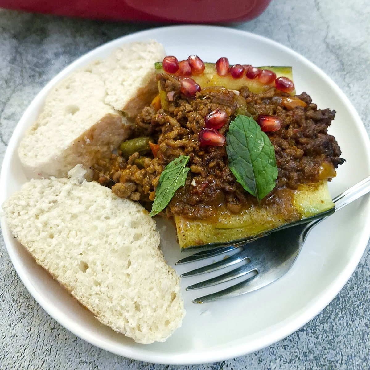 A slice of lamb keema stuffed marrow on a white plate accompanied by 2 slices of crusty bread.