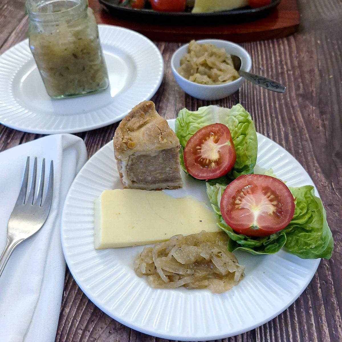 A small white plate piled with a pork pie, lettuce and tomato, cheese and a spoonful of onion marmalade.